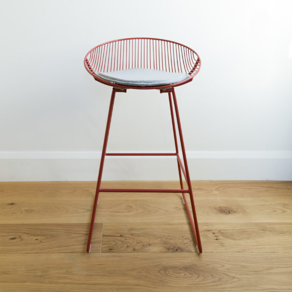 A grey, felted wool, round stool pad sits on a red wire kitchen barstool