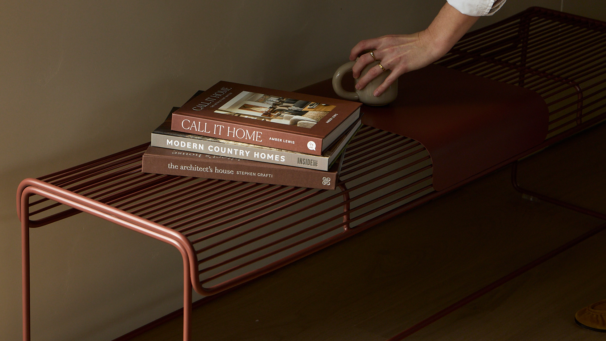 Benmore Bench in Chocolate Red with books and a ceramic mug, showcasing the powdercoated steel wire bench with a moveable metal plate.